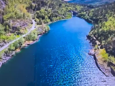 Aerial shot of a fjord or long lake surrounded by forested hills, likely in Norway. The water is a vibrant blue with clear visibility and ripple patterns. A road follows the shoreline on the left. A waterfall is visible in the distance at the far end of the lake. The image does not appear to be in Basingstoke and Deane, United Kingdom.