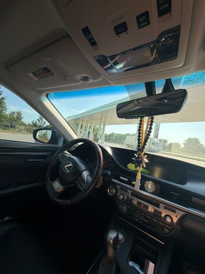 The media file shows the interior of a car, likely a Lexus, as indicated by the logo on the steering wheel. The view is from the passenger seat, looking towards the driver's side and the front windshield. The time of day appears to be daytime, with bright sunlight streaming through the windshield, suggesting fair weather.
Dominant objects in the interior include the steering wheel, the dashboard with various buttons and controls, a rear-view mirror, and the overhead console with controls for "CLOSE," "DOOR," and "SOS" (Safety Connect). There are also decorative elements hanging from the rear-view mirror, including beaded strings and a star-shaped ornament. The sun visor is flipped down, displaying warning text in both English and French: "AIR BAG WARNING FLIP VISOR OVER" and "COUSSIN GONFLABLE AVERTISSEMENT REPLIER LE PARESOLEIL."
Through the windshield, the scene outside the car is visible. It appears to be a gas station with canopies and fuel pumps. Trees and greenery are also visible in the background, suggesting an outdoor, possibly rural or suburban setting. A white truck is seen in the distance.
The overall impression is of a car parked at a gas station on a sunny day. There are no people visible in the image. The focus is on the car's interior and the immediate exterior environment.