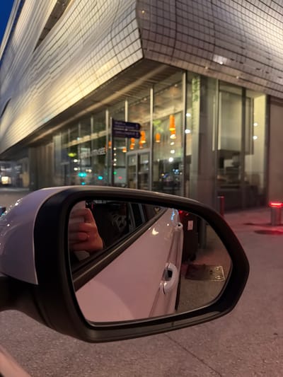 The image shows a view from inside a vehicle, looking at the side-view mirror. The mirror reflects a person's hand with a ring on their finger, and a portion of the car's interior. Outside the vehicle, a modern building with a distinctive corrugated facade is visible. The building features large glass windows and is illuminated, suggesting it is evening or nighttime. Orange pendant lights are visible inside the building through the windows. A street sign with purple and white text is also present outside. Red bollards with illuminated tops are seen along the sidewalk. The location is identified as being in Nîmes, France.
