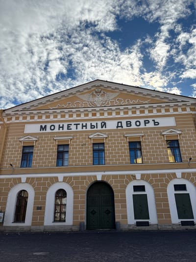 The image shows a building with the inscription "МОНЕТНЫЙ ДВОРЪ" in large, dark letters on a white horizontal panel. This inscription translates to "Mint Yard" or "Mints Court." The building is painted in a warm, yellowish-orange color with white trim, and it has a pediment at the top featuring a relief of a double-headed eagle with crowns, wings spread. Below the inscription are several arched windows with dark bars, and a large, ornate green double door in the center of the facade. The building's facade is constructed of bricks, with white lines outlining the brickwork. The ground in front of the building is paved with dark cobblestones. The sky above is blue with scattered white clouds.
This building is the Saint Petersburg Mint (Monetny Dvor), a historic mint located in Saint Petersburg, Russia. It is one of the oldest mints in Russia, founded in 1724.