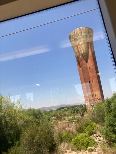 The image captures a reflection of a tall, brick water tower against a clear blue sky, seen through a window. The water tower appears old, with a wider, curved base that tapers into a cylindrical shaft. The bricks are a reddish-brown color, and there are visible windows or openings along the shaft. Text is spray-painted on the tower, with the visible portion appearing to read "ISC." to the right of the tower and possibly part of another word.
The scene outside the window is a landscape with lush green trees and vegetation, suggesting a rural or semi-rural setting. There's a wire fence visible in the background, and the terrain beyond seems somewhat hilly. The bright sky and the clear visibility of the landscape indicate it is daytime with good weather, likely sunny. The reflection on the window suggests the photo was taken from inside a moving vehicle, such as a train or car, as indicated by the slight tilt and the streaks on the glass. The overall mood is neutral, focusing on the architectural structure and the natural surroundings. The location context of El Vendrell, Spain, would place this scene in a Mediterranean landscape.