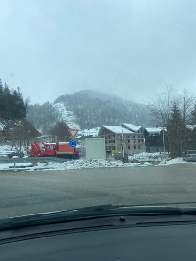 The image, taken from inside a vehicle, shows a snowy mountain scene. In the foreground, a gray road is visible. To the right, a portion of the vehicle's interior is visible. On the left side of the road, there is snow covering the ground. Further into the scene, there is a red snowplow with a blue and white roundabout sign and a yield sign behind it. In the background, there is a mountain covered in snow and trees, obscured by fog. A building is visible on the right side of the frame. The location is Katun Bačko brdo, Montenegro.