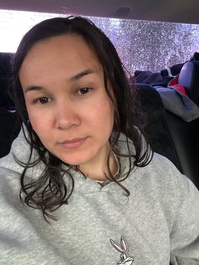 A young woman with dark hair is seated in what appears to be a car. She is wearing a grey sweatshirt featuring a Bugs Bunny design. The car window beside her is covered in water droplets, suggesting rain. The background is indistinct but hints at urban features possibly located in Uray, Russia. Other clothing items are visible in the background, likely on the seat behind her.