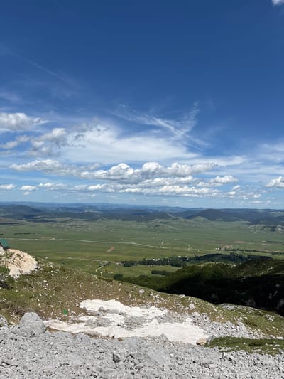 A wide-angle, high-angle shot captures a vast, verdant valley under a bright blue sky dotted with fluffy white clouds and wispy cirrus formations. In the foreground, a rocky, barren slope is sparsely covered with dry grass and patches of melting snow. A small, green-roofed building is visible on the left side of the foreground slope. The midground is dominated by rolling green hills and a sprawling valley floor, crisscrossed by a winding road and dotted with small clusters of buildings and cultivated fields. The background consists of layered, hazy mountain ranges fading into the distance under the expansive sky. The scene appears to be taken from a mountaintop overlooking a rural landscape in or near Pašina Voda, Montenegro.