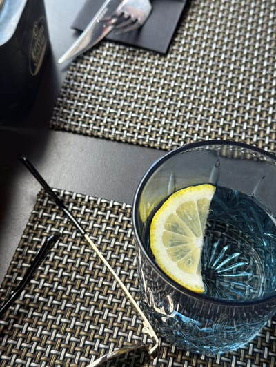 This is a close-up, overhead shot of a table setting with a woven placemat, a glass of water with a lemon slice, and a pair of eyeglasses. The glass is clear with a blue tint and has a faceted design. The lemon slice is bright yellow and positioned in the upper right quadrant of the glass. The eyeglasses have thin, gold-colored arms and dark frames. In the lower left of the image, a silver fork rests on a black napkin. The background is a textured, woven placemat with alternating brown, white, and grey threads. The lighting suggests it could be daytime, possibly indoors or on a shaded patio, given the soft light. The setting appears to be a restaurant or cafe, indicated by the cutlery and placemat. No discernible text is visible in the image.