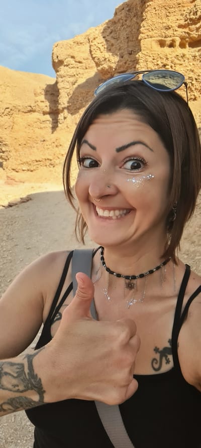 The image shows a young woman with shoulder-length dark hair. She is smiling broadly and giving a thumbs-up. She has light skin and is wearing makeup, including silver glitter near her eyes. She has a nose piercing. Her sunglasses are resting on her head. She is wearing a black tank top with spaghetti straps and a black beaded necklace with silver chains hanging from it. She has tattoos on her collarbone and wrist. She is standing outside near a tan-colored rock formation or wall, with blue sky visible in the background.
Given the presence of the tan-colored rock formation and the sky in the background, it is difficult to determine a more precise location within the Hevel Eilot Regional Council area without additional contextual cues in the photograph.