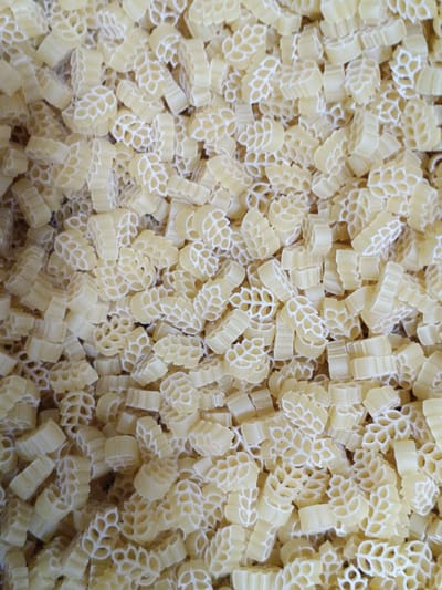 A close-up, overhead shot reveals a textured surface composed of numerous small, light yellow pasta pieces. The pasta is shaped like tiny wheat stalks, with a hollow, intricate design within each piece. The pieces are densely packed, creating a uniform and slightly varied texture due to their overlapping arrangement. The lighting is even, casting no distinct shadows and highlighting the pale color and detailed shapes of the pasta. There is no visible context of location, such as a city in Martuni, Armenia or any other environmental features. The image exclusively focuses on the pile of pasta.