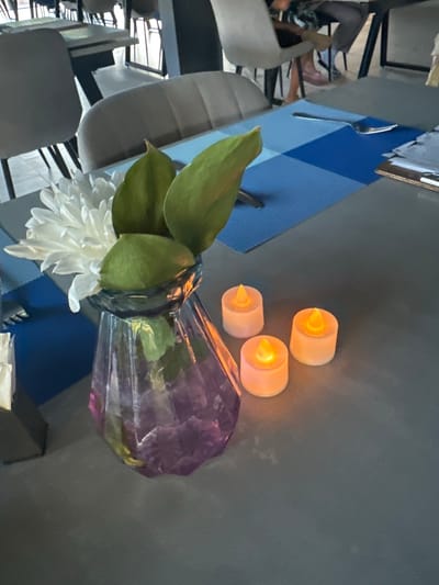 The image depicts a table setting in what appears to be a restaurant. In the foreground, a purple glass vase holds a white chrysanthemum and some green foliage. Three small, battery-operated candles with orange flames are arranged to the right of the vase. The table is covered with a gray surface, and a blue checkered placemat is visible behind the vase. In the background, several chairs and other tables are present, suggesting an indoor dining area. The overall scene is well-lit, with the candles providing a warm glow. There is no visible indication of the specific location being in Даниловка (алтайский), Russia.