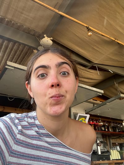 A young woman is depicted in a close-up selfie, making a direct eye-level gaze with a wide-eyed, pouting facial expression. She has dark hair, prominent eyebrows, and is wearing light-colored drop earrings and a striped, off-the-shoulder top featuring white, red, and blue/grey stripes. Her skin shows a slight reddish flush around the nose and cheeks. The scene is located within the Megilot Yam HaMelakh Regional Council, Palestinian Territory. The background reveals a semi-enclosed outdoor setting, likely a cafe or bar. An overhead canopy of tan fabric is visible, supported by wooden beams, with a section of corrugated metal or wood paneling above it. An exposed incandescent light bulb hangs from a bamboo-like support to the upper right. Above the woman's head, a small white object, possibly a light fixture, is partially visible. To the right, a bar area is present, featuring inverted wine glasses on a rack, various bottles of beverages on shelves, a coffee machine or tap system, and a stack of small cups on a wooden counter. A colorful framed item hangs on the wall behind the bar.