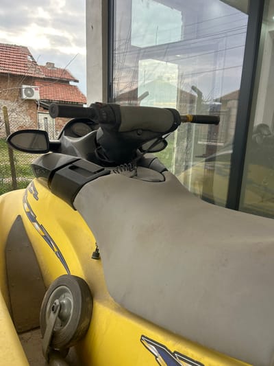 A yellow personal watercraft, coated in a layer of dust, is parked under a covered structure in Dolni Tsibar, Bulgaria. The vessel features a light gray seat and dark gray handlebars, equipped with side mirrors and control mechanisms, including a coiled lanyard. Blue and white graphic decals are present on the yellow hull. A small gray wheel assembly is visible positioned against the left side of the jet ski. Directly behind the personal watercraft, a large glass window reflects portions of the exterior environment. Through this window, an overcast sky is visible above a residential building constructed of reddish-brown brick with a matching tiled roof. A white outdoor air conditioning unit is mounted on the building's exterior wall. Bare tree branches and a wire fence are also discernible in the background. No individuals are present, and no actions are occurring.