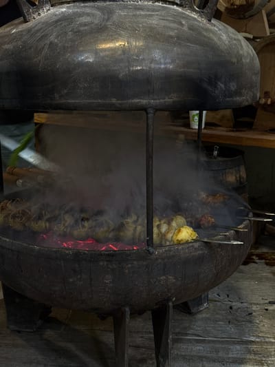 A close-up, medium shot captures a large, weathered, black cauldron with a domed lid suspended over glowing red coals. Inside the cauldron, several skewers of food are being grilled. The food on the left appears to be whole, rounded items, possibly potatoes or onions, arranged in a line. To the right, there are skewers with chunks of meat, also being cooked over the coals, and enveloped in thick white smoke that billows upwards. Wooden handles of some utensils or skewers are visible near the top left of the cauldron. The cauldron is supported by three dark, metallic legs. In the background, a wooden shelf holds barrels and a white paper cup with a green logo. The flooring beneath the cauldron is made of dark wood planks. This scene is set in Sisian, Armenia, at a location that appears to be an outdoor food stall or preparation area.