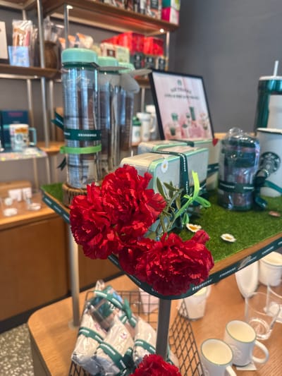 The image shows a display of Starbucks merchandise in a retail setting, likely a Starbucks store in Gimpo-si, South Korea. The merchandise includes tumblers, coffee beans in cylindrical containers, gift boxes, mugs, and other packaged goods. The Starbucks logo is visible on many of the items and ribbons. Red artificial flowers are arranged in the foreground. The display is set on a wooden table. A shelf with other merchandise is visible in the background. The lighting is bright, indicating an indoor environment.