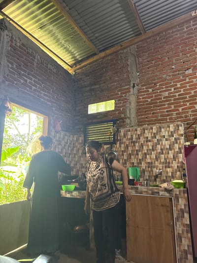 In Babahjurong, Indonesia, two women are in a rustic kitchen. The walls are constructed of red brick, and a corrugated metal roof with visible wooden supports is overhead. Natural light streams in through a large open window on the left, revealing lush greenery outside. A smaller, frosted glass window is present on the right wall.

One woman, wearing a dark, long-sleeved dress, stands by the window, facing away from the camera. The other woman, in a patterned blouse and dark pants, is near the kitchen counter, which is tiled with a mosaic of brown, beige, and white squares. She appears to be interacting with the counter or cabinets.

On the counter, there is a green plastic pitcher, a faucet, a bottle of water, and two green bowls. A stove with a cooking pot is visible behind the woman in the patterned blouse. A pink refrigerator is partially visible on the far right. The overall environment suggests a simple, functional living space.