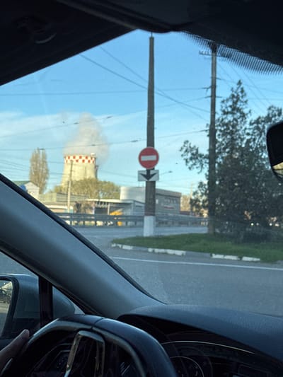 The image captures a view from the interior of a vehicle, looking out through the windshield. The scene depicts a roadside industrial area, likely during the daytime under a clear sky.
**Subjects:** The foreground shows parts of the car's interior, including the textured dashboard on the right, a portion of the steering wheel, an A-pillar, and a side mirror. The windshield shows reflections of the car's interior. Outside the vehicle, there is a paved road to the right, bordered by a grassy verge. A tall utility pole stands prominently in the middle-left, supporting electrical wires. Beyond the pole, trees are visible, some with sparse foliage. The most prominent structure in the background is a large industrial tower, possibly a cooling tower or smokestack, characterized by a distinctive red and white checkerboard pattern. It emits a substantial plume of white smoke or steam into the blue sky.
**Activity or Event:** The image suggests the viewer is inside a car, either driving or parked, observing the industrial landscape.
**Notable Details:** The time of day appears to be daytime, with clear skies, though the sun's position is not directly visible. The large industrial tower with its characteristic checkerboard pattern and the emitted smoke strongly indicate a power plant or heavy industrial facility. This detail aligns with the provided location context of Gresovsky, Ukraine, known for a thermal power plant.
**Visible Text:** Attached to the utility pole are two traffic signs. One is a standard "No Entry" sign, a red circle with a horizontal white bar. Below it, a white square sign with a black arrow points to the right, likely indicating a mandatory direction or a detour.
