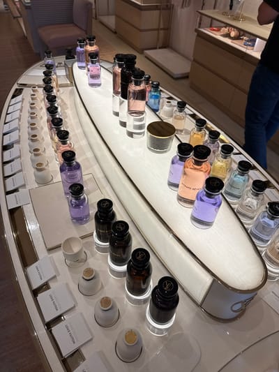 This media file captures a close-up view of a luxury perfume display at a Louis Vuitton store, likely in Oak Brook, United States of America. Numerous Louis Vuitton perfume bottles in various vibrant colors, including light blue, purple, pink, amber, and dark brown, are neatly arranged on a sleek, curved, and illuminated white counter. Each bottle features a black cap adorned with the Louis Vuitton logo.

In front of several bottles, small white ceramic scent testers or cups are positioned, inviting customers to experience the fragrances. Accompanying these testers are small white informational cards, each displaying the perfume's name and "LOUIS VUITTON." A person wearing blue jeans is partially visible in the upper left, standing behind the counter, suggesting staff presence or another customer. The overall scene conveys an upscale and meticulously presented retail environment.

Visible text includes product names such as "ATTRAPE-RÊVES," "IMAGINATION," "OMBRE NOMADE," and "LES SABLES ROSES," which appear on both the bottles and their corresponding informational cards. "LOUIS VUITTON" is also prominently displayed on almost all cards. Other partially visible texts on bottles include "L'IMMENSITÉ" and a card for "FLEUR DU DÉSERT." The bright, even lighting emphasizes the products and the luxurious setting of the store.