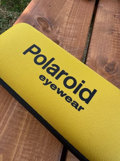 A close-up, high-angle shot shows a yellow hard-shell glasses case with "Polaroid eyewear" printed in black text on its lid. The case is placed on a weathered wooden surface, possibly a picnic table or bench. The wood grain is prominent, with visible knots and lines. In the background, out of focus, is green grass and more wooden planks. The scene is outdoors, and the lighting suggests it is daytime. No people are visible. The location is Zgorigrad, Bulgaria.