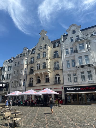 This is a vertical, outdoor photograph taken on a bright, sunny day with a clear blue sky and wispy clouds. The scene depicts a European streetscape with ornate, multi-story buildings lining one side and a cobblestone plaza on the other. Several people are visible.

In the foreground, a woman with dark hair, wearing a plaid shirt and denim shorts, walks away from the camera, carrying a backpack. Several empty outdoor tables and chairs are set up on the cobblestone plaza. Further back, under white umbrellas with "Pizza Hut" branding, more tables are occupied by patrons dining and socializing. The buildings are characterized by their intricate architectural details, including decorative facades, numerous windows with arched tops, and balconies.

Visible text includes "Pharmacie" on one building and "Münster apotheke" on another. Below the "Münster apotheke" sign, a store with a glass facade displays various products. A black sign with "KS" in white letters is also visible at the bottom edge of the frame. The overall impression is a lively street scene in a European city, likely during daylight hours given the bright sunshine.