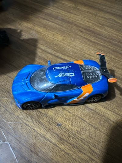 A blue toy race car with orange accents is positioned on a wooden surface. The car has a prominent spoiler on its rear, with an orange tip. The car's body is adorned with white decals. One decal features a stylized graphic and the number "1150", with smaller text that appears to read "BEHANCE STUDIO". Another decal on the side shows a white "1". The wheels are black with silver rims. The wooden surface has a visible grain pattern and appears to be a tabletop or floor. The lighting suggests an indoor setting, possibly with artificial light casting shadows. There are no people or animals visible in the image. The overall scene is a static, still life arrangement of the toy car.