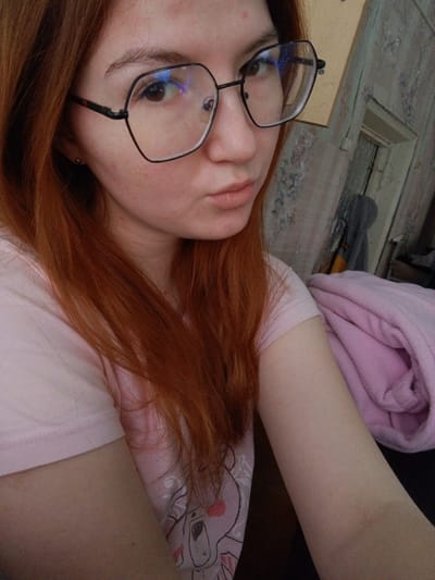A young adult female, located indoors in the city of Старый Шагирт, Russia, is depicted in a close-up, high-angle photograph. She has long, reddish-brown hair and light skin with minor facial blemishes on her forehead and cheeks. She wears large, black-framed hexagonal eyeglasses that exhibit blue reflections on the lenses. Her lips are slightly pursed, forming a pout, and her gaze is directed towards the camera. She is wearing a light pink short-sleeved top with a faint white and pink cartoon graphic visible on the right side of her torso. The background on the left and center consists of a wall covered in faded, patterned wallpaper. To the right, a rolled pink textile object, likely a blanket or towel, rests on a dark surface. Beyond this, a light-colored, weathered wooden doorframe is visible, with a dark, indistinct item draped or hanging in the doorway opening.