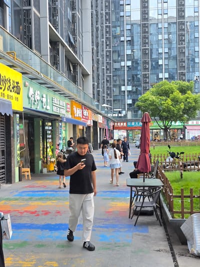 This image captures a street scene in Linping District, China, with a focus on people and businesses lining a pedestrian walkway. The setting appears to be urban, with a modern building with numerous windows and air conditioning units dominating the background on the left. The foreground features a walkway with colorful chalk art, suggesting a lively and possibly artistic atmosphere. Several people are visible, including a man in a black shirt and light pants looking at his phone, and a group of women and children walking by.

The businesses along the street include what appear to be restaurants or food stalls, indicated by the visible signage with Chinese characters. One sign explicitly reads "汤面盖面" which translates to "Soup Noodles and Covered Noodles," and another mentions "鲜活超市" meaning "Fresh and Live Supermarket." There is also a visible sign for "手机维修" (mobile phone repair) and "康大药房" (Kangda Pharmacy), suggesting a mix of retail and service establishments. A red umbrella is positioned near a table and chairs, hinting at outdoor seating arrangements for a restaurant or cafe.

The time of day appears to be daytime, likely late afternoon, given the lighting. The weather seems pleasant. The overall impression is a bustling commercial street with a blend of modern architecture and local businesses, enhanced by the vibrant street art.