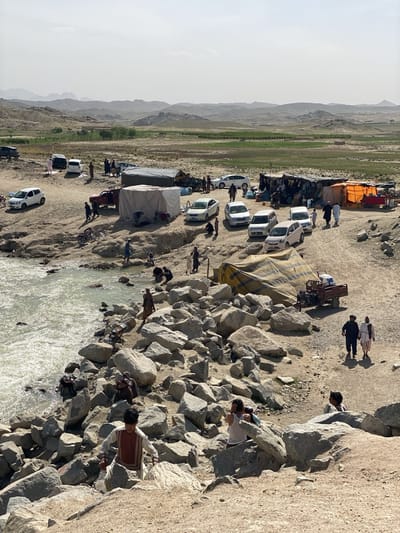 The image shows a scene in a rural area of Kim, Afghanistan. In the background, there are arid hills and a flat green field. Several cars and tents are scattered across a flat, rocky landscape. People are present, some gathered near the tents and cars, others near a river with rocky banks. Some individuals appear to be wading or standing in the water. One person is climbing the large rocks at the bottom of the frame. Other objects include motorcycles, water containers and farm equipment.