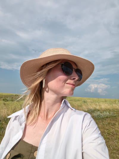 A woman wearing a straw hat and sunglasses stands in a grassy field under a cloudy sky. She is looking to the right of the frame, and her blonde hair is blowing in the wind. She is wearing a white collared shirt and a green outfit underneath. The background is a blurred expanse of grass and a distant horizon. The scene is set in an outdoor, natural environment, possibly a field or meadow. No city or specific location details are visible in the image.