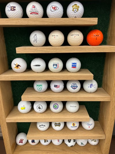 An indoor, eye-level, medium shot shows a wall-mounted, multi-tiered display case filled with golf balls. The display case is constructed of light-colored wood with three visible shelves. The background behind the shelves is a dark green, textured material, possibly felt or astroturf. The shelves are filled with golf balls, each displaying a logo or text. The top shelf holds four golf balls. From left to right, the first has a white ghost with a purple 'E'. The second is white with a red logo and the text "MULES NATIONAL GOLF CLUB PERTLE SPRINGS". The third is white with a red outline of a KC Chiefs logo. The fourth is white with a yellow and black tiger face logo. The second shelf from the top also has four golf balls. From left to right, the first is white with a white Michelin Man character. The second is white with the text "RAM 3D" and the number 4. The third is white with black lettering that appears to be "XOXXXXXX" and the number 7. The fourth golf ball is bright orange with "PING" and the number 1. The third shelf from the top contains four golf balls. From left to right, the first has a gold eagle crest logo. The second has a black depiction of a bullet. The third has a blue and red shield logo with text that says "AMERICAN SHIELD INSURANCE". The fourth has a blue and red logo with "SHELTER INSURANCE". The fourth shelf from the top has four golf balls. From left to right, the first has a green shamrock logo and "ST. PATRICK'S IRISH OPEN". The second has an American flag design. The third has a circular logo with a starburst and "PATRICK'S GOLF DAY". The fourth has a white skull and crossbones logo. The fifth shelf from the top has four golf balls. From left to right, the first has a yellow Masters logo. The second has a black silhouette of a person and a tree, with "COUNTRY CLUB OF MISSOURI 1971". The third has a circular logo with a green shamrock and "Butler Creek Country Club". The fourth has a black and white "Good Goods" logo. The sixth shelf from the top has four golf balls. From left to right, the first has a black logo with "Jim Beam BLACK". The second has "Bushmills IRISH WHISKEY". The third has a blue and red Foster's