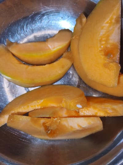 The image shows several slices of cantaloupe melon arranged in a metallic bowl. The cantaloupe is a vibrant orange color with visible seeds and pulp in some slices. The bowl has a slightly textured, reflective surface, suggesting it is made of stainless steel. The lighting appears to be artificial, casting shadows and highlighting the sheen of the bowl and the juicy texture of the fruit. The scene is a close-up shot, focusing entirely on the fruit and its container. There are no people or text visible in the image. The setting is likely a kitchen or dining area, given the presence of a food item and a bowl. The overall impression is one of freshness and simple food preparation.