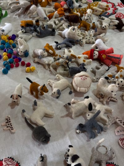 The image displays a collection of handmade felt animal figures arranged on a white linen surface. The figures are primarily small and appear to be crafted from wool. They depict a variety of animals including dogs, cats, rabbits, and possibly a mouse, rendered in a simplified, stylized manner. Some figures are standing, while others are lying down or in motion. There are also some loose felt balls and beads in bright colors scattered among the figures, as well as a doll dressed in red and orange clothing. The lighting suggests an indoor setting, likely a market or craft display. The image does not contain any specific landmarks or contextual clues that would allow for a precise location within Кызыл-Туу, Kyrgyzstan. It is a close-up shot focusing on the texture and detail of the felt creations.