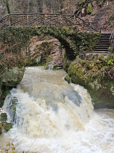 The image depicts a natural landscape in Waldbillig, Luxembourg, dominated by a fast-flowing, light brown river with multiple cascades. The river exhibits significant whitewater and foam as it flows over and around moss-covered rocks. An arched stone bridge spans the river, its structure heavily covered in green moss and ferns. The bridge deck features a rustic wooden handrail constructed from an 'X'-patterned arrangement of interlocking branches. To the right of the bridge, a set of wooden steps with a similar wooden handrail ascends a steep, tree-covered embankment. The surrounding environment consists of a densely wooded area with predominantly bare deciduous trees and numerous large, moss-covered boulders. A small, brown informational sign is affixed to a tree or post on the hillside above the stairs. No human subjects are visible.