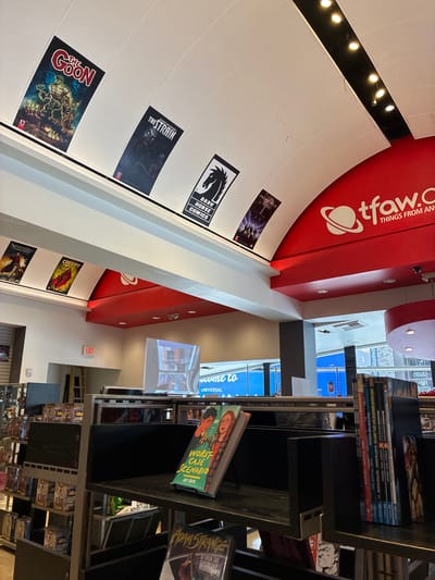 The image shows the interior of a comic book store. Various comic book covers and posters are displayed on the white, arched ceiling. In the foreground, there are bookshelves filled with comic books and graphic novels. One book prominently displayed on a shelf in the foreground is titled "WORST CASE SCENARIO" by Ray Stieve. To the right, a stack of comic books with various titles, including "AQUAMAN," are visible. A red sign with a white planet logo and "tfaw.com THINGS FROM ANOTHER WORLD" is mounted on a curved red structure above the bookshelves. An "EXIT" sign is visible on a light-colored wall in the background, to the left of a large screen displaying a blue graphic. The store appears to be well-lit with artificial lighting.