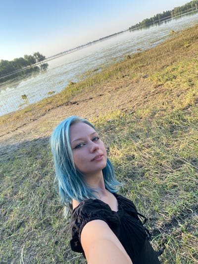 A young person with blue hair lies on their side on dry grass. They are wearing a black garment. The person's face is turned towards the camera, and they have eye makeup on. The background shows a body of water and a clear sky, suggesting it is daytime and the weather is fair. The overall scene is outdoors, with the water possibly indicating a lake or river. The image is taken at a low angle, with the person dominating the foreground. The setting appears to be a natural, open landscape. No visible text is present in the image.