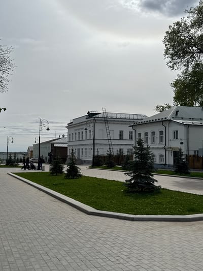 The image depicts a scene in Arzamas, Russia, showing a white, multi-story building with a grey roof. A metal ladder extends from the ground to the roof of the building. The architecture suggests a possibly historical or administrative building. Adjacent to the building is a strip of green lawn bordered by a grey paved walkway. Streetlights are visible along the walkway, extending into the distance. In the distance, there are hints of other buildings and infrastructure. The sky is overcast. The scene includes individuals that seem to be sitting on a bench alongside the street.