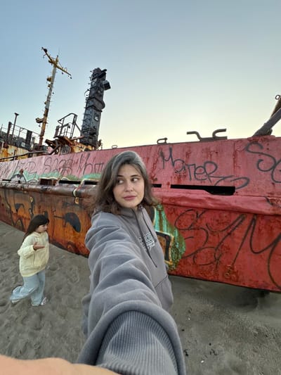 Two females, an adult and a child, are on a sandy beach in Teriberka, Russia, in front of a large, rusted shipwreck covered in graffiti. The adult, in the foreground, is taking a selfie with a gray hoodie. The child, in a yellow jacket and blue pants, stands to the left, gesturing with her right hand. The shipwreck dominates the background, its red hull and rusty metal structures weathered by the elements. The sky above is a pale blue.