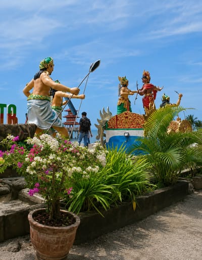 The image captures a scenic outdoor view at the Kota Pari attraction in Indonesia, under a bright blue sky with scattered clouds. Dominating the foreground is a vibrant bougainvillea plant in a terracotta pot, adorned with pink and white blossoms, nestled amongst lush green foliage.
In the background, a series of colorful statues depicting figures from what appears to be a mythological or historical scene are prominently displayed. One statue, a warrior in a green headdress and blue skirt, holds two spears, aiming them towards the right. To the right, two figures in ornate attire engage in what looks like a ceremonial gesture or perhaps a duel, one holding a bow and arrow. Another figure is visible further to the right, raising an arm.
A person wearing a black backpack and blue jeans walks away from the camera, past the statues, towards the sea visible in the distance. Signs with large green letters, partially obscured, are also present to the left, suggesting the name of the location. The overall setting is well-maintained, with stone pathways and decorative planters contributing to the attractive presentation of the Kota Pari attraction.