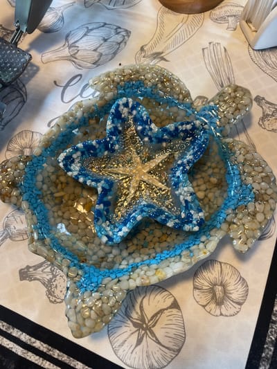 A close-up, top-down shot shows a resin art piece in the shape of a turtle, filled with small, smooth pebbles and a blue and white starfish ornament in the center. The turtle-shaped piece is on a tablecloth with a printed design of various vegetables and calligraphy. The scene is indoors, likely in a home, with no indication of the city of Турово, Russia.