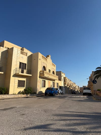 This is a ground-level, eye-level, eye-level, medium shot of a street in Xagħra, Malta. The sky is clear and blue, with no clouds visible. The street is paved with asphalt and has white markings. There are several cars parked on the side of the street, including a blue hatchback, a white car, and a white pickup truck. On the left side of the street, there is a yellow building with stone accents and balconies. On the right side of the street, there are more buildings, also with stone accents and balconies. A palm tree is visible on the far right. The lighting suggests it is late afternoon or early morning, with long shadows cast across the street. The overall impression is a quiet residential street in a Mediterranean town.