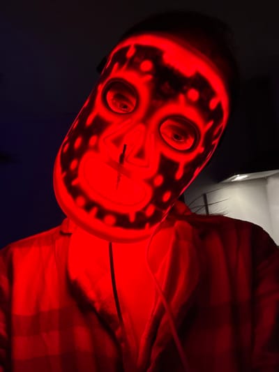 The image shows a person wearing a red mask with illuminated facial features and dot patterns. The person is wearing a red plaid shirt. The background is dark, with a glimpse of a light-colored ceiling and wall. The location (Kranj, Slovenia) is not evident in the image.