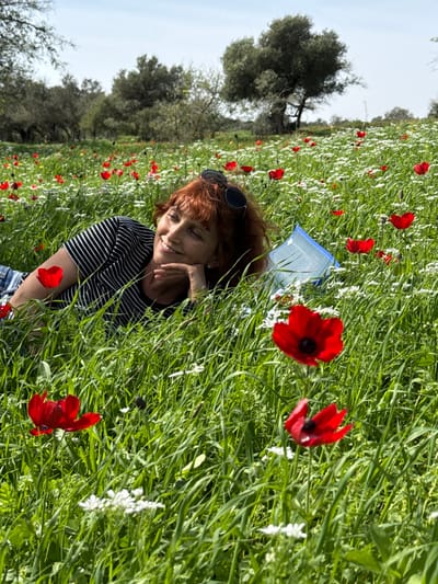 A solitary woman with reddish-brown hair is depicted lying on her left side in an expansive field. She wears a black and white striped short-sleeved top, with sunglasses resting on her head. Her left arm is bent, supporting her head which rests on her left hand. Her eyes are closed, and her mouth exhibits a slight smile. The field is densely covered with vibrant green grass, punctuated by numerous prominent red anemone flowers and smaller white wildflowers. A segment of a light blue and white patterned mat is visible partially beneath her. In the background, several trees with sparse foliage stand against a bright blue sky with minimal cloud cover. This natural open field is situated within or bordering Modiin-Maccabim-Reut, Israel, reflecting the typical springtime flora of the region.