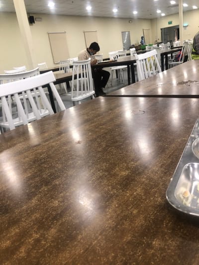An eye-level shot in a brightly lit cafeteria features a long, dark wood table taking up most of the frame. It has chairs on either side. A person wearing a beige shirt and a mask sits at the table, legs stretched out under a chair. A tray with food sits on the corner of the table. White chairs extend toward the back, leading to a distant background that shows a window. The background also shows blank, beige walls.