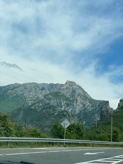 A roadside view in Donji Jasenjani, Bosnia and Herzegovina, presents a gray asphalt roadway with white directional arrows adjacent to a metal guardrail. A diamond-shaped road sign stands amidst green foliage beside the road. A towering, rugged mountain with exposed gray rock faces and patches of green vegetation dominates the scene, juxtaposed against a blue sky with scattered clouds. Overhead power lines stretch across the landscape.