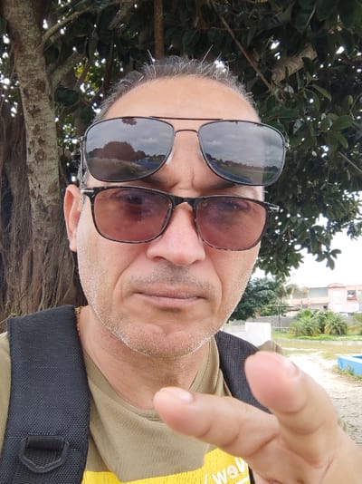 The image features a man taking a selfie outdoors, presented in a portrait orientation but originally captured sideways (rotated 90 degrees clockwise). The man, likely middle-aged or older, is the central focus of the frame. He has fair skin, a light, stubble-like beard or mustache, and is wearing sunglasses with clip-on dark lenses over what appear to be regular eyeglasses. His hair on the sides of his head is short and grey, while the top of his head appears bald or very closely shaven. He has a neutral expression, with his mouth closed.
He is wearing a light tan or olive-green short-sleeved shirt. A black strap, possibly from a backpack or shoulder bag, crosses his left shoulder. A thin gold-colored chain is visible around his neck. His right hand is raised towards the camera in the foreground, with his palm facing outwards and fingers slightly spread, perhaps in a greeting gesture.
The background behind him is split between natural elements and a distant urban setting. To his right, there are large tree branches with visible bark texture and abundant green leaves, suggesting a lush outdoor environment. To his left and in the lower background, several residential buildings or houses with varying rooflines are visible. In front of these buildings, there is an open, somewhat overgrown area, possibly a field or undeveloped land, with some smaller green plants and what appears to be a distinct blue linear structure in the foreground, possibly a border or part of a small structure.
The lighting is bright and natural, indicating it is daytime. Reflections in his sunglasses show a bright sky with some clouds, along with reflections of the surrounding trees and possibly distant structures, further confirming the outdoor setting. On his inner shirt, visible through the lighter outer shirt, some partial text in yellow is visible, reading "we GM" or "we're GM", with the letters "we" and "GM" clearly distinguishable.