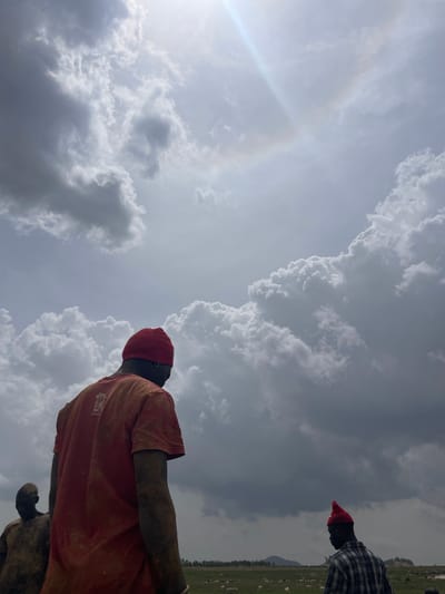 A medium shot, taken from a low angle and rotated counterclockwise, shows two individuals under a cloudy sky. The primary subject, closer to the camera, wears an orange t-shirt, a red beanie, and appears to be covered in dirt or mud. A secondary individual, positioned to the right and further away, wears a dark, plaid shirt and a red head covering. The sky is overcast with prominent cumulonimbus clouds and a visible sun halo. In the background, a field extends towards the horizon, populated with small, indistinct white objects, potentially livestock. The location is Maiyanga, Nigeria.
