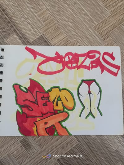 The image is a close-up, top-down shot of a sketchbook page featuring graffiti-style artwork. The sketchbook is lying on a light brown wooden floor with a parquet pattern.
On the sketchbook page, there are two distinct graffiti pieces. The top piece is large, red lettering with a yellow outline, appearing to spell "DOZS". The lettering is bold and stylized, with thick strokes and sharp angles.
Below the red lettering, there is another graffiti piece primarily in red and orange with a green outline, appearing to spell "SERR". This piece is more intricate and flame-like in its design. To the right of this, there is a stylized drawing of a pair of buttocks wearing red underwear, outlined in green and with yellow shading.
The overall impression is that of street art or personal artistic expression within a sketchbook. The background of the wooden floor suggests an indoor setting, possibly a home or studio. The "Shot on realme 8" watermark in the bottom right corner indicates the image was taken with a mobile phone. There is no information in the image that places it in Shelekhov, Russia.