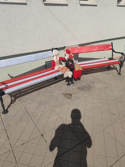 In the city of Nevestino, Bulgaria, a park bench is positioned on grey concrete paving tiles against a light-colored building wall with two visible windows. The bench features black wrought iron supports; its slats are wrapped in alternating red and white material, with the backrest showing a white section on the left and a red section on the right. Two stylized figures, constructed from woven fabric or yarn, are seated on the bench. The figure on the left presents a white head, a red torso, and beige legs with red crisscross bindings and white tassels. The figure on the right is predominantly red, characterized by a tasseled lower body and a small, rectangular brown object resting on its lap. Strong overhead sunlight casts a distinct human shadow, indicative of the photographer, onto the paved foreground.