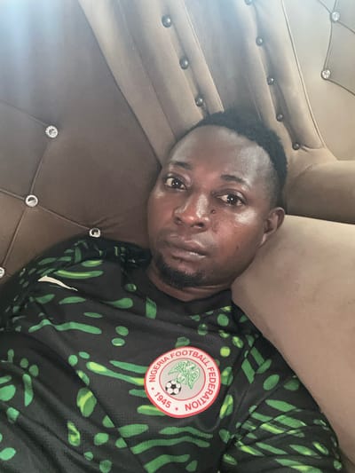 A man with dark skin and short black hair, wearing a black and green patterned jersey with a crest that reads "Nigeria Football Federation 1945", lies on a brown tufted couch with crystal embellishments, in Gidan Waziri, Nigeria. The man is looking directly at the camera with a neutral expression. The couch appears to be upholstered in a velvet-like material and has a prominent crease in the center. The lighting suggests an indoor setting.