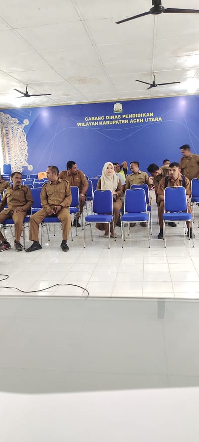 In Meunyematangubi, Indonesia, a group of people are seated on blue chairs in what appears to be an indoor meeting or event space. The room has a high ceiling with white tiles and two black ceiling fans. A large blue banner with yellow text hangs on the back wall, displaying "CABANG DINAS PENDIDIKAN WILAYAH KABUPATEN ACEH UTARA". A symbol with the text "PANCASILA" is also visible on the banner. The individuals are primarily men, all dressed in tan-colored uniforms, and one woman wearing a beige headscarf and tan uniform. Some are looking towards the camera, while others are engaged in conversation or looking elsewhere. A black cable runs across the floor in the foreground. The overall impression is a formal gathering in a public facility.