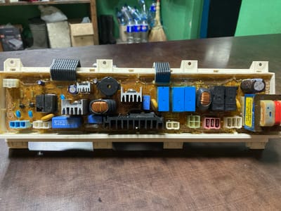 The image shows a close-up, eye-level view of a dismantled electronic circuit board, likely from a household appliance such as a washing machine. The circuit board is laid flat on a dark, wooden surface, possibly a table. The board itself is rectangular and densely populated with various electronic components, including resistors, capacitors, relays, transformers, and integrated circuits. Some components are covered in a yellow, translucent substance, suggesting protective coating or adhesive. The background is out of focus, but appears to be an indoor setting with shelves and objects, including bottles and cleaning supplies, visible in the distance. The lighting is artificial and casts a slight shadow. There are no people or animals present in the image. The location is not specified within the image, therefore its presence in Ukrainka, Ukraine, cannot be confirmed.