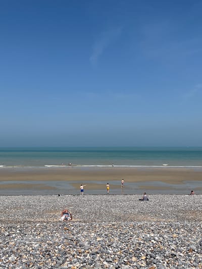 The image captures a view of the pebble beach and the English Channel in Dieppe, France. The sky is a clear, bright blue with a faint haze on the horizon. The sea is a tranquil turquoise, with small waves gently lapping at the shore.
The foreground is dominated by a vast expanse of pebbles, ranging in color from white and grey to darker hues. Several people are scattered across this pebble beach, either relaxing or sunbathing. In the middle ground, the wet sand of the receding tide creates reflective pools. A few individuals are walking or playing in the shallow water. Further out, more people are visible near the water's edge. The overall impression is one of a peaceful, sunny day at the beach.