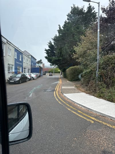 The image depicts a street scene in Courtown, Ireland. A blue and white building labeled "Courtown Hotel" is visible on the left side of the road, with several cars parked in front of it. The road itself is asphalt with double yellow lines marking the center. A sidewalk runs alongside the road, bordered by a hedge and trees. A streetlamp stands on the right side of the road, and a single black car is driving away in the distance. The weather appears overcast. The reflection in the side mirror of a vehicle suggests the photo was taken from inside a car.
